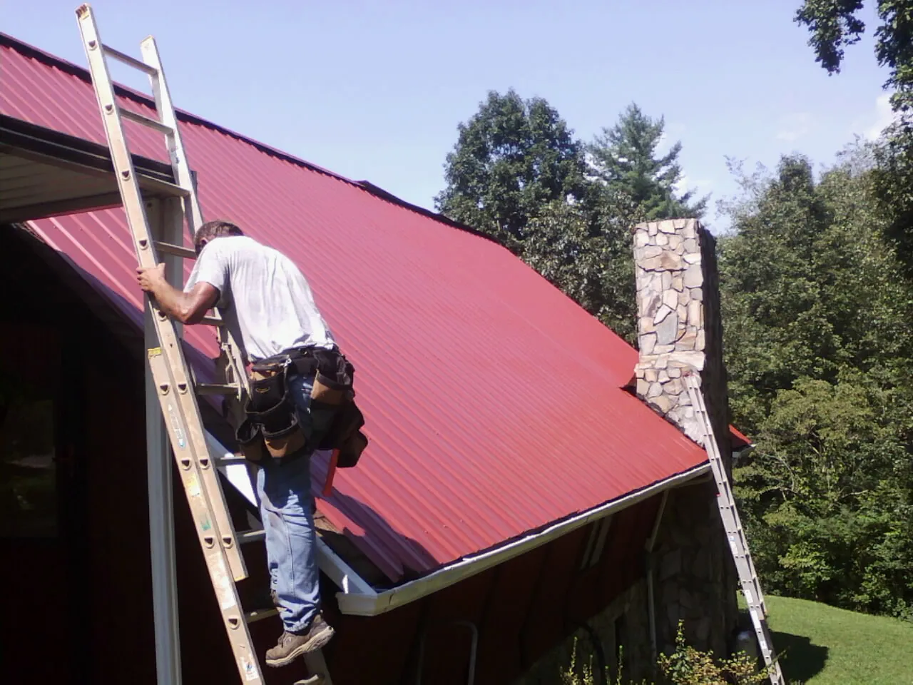 Roofer on a ladder beside a red metal roof and stone chimney — Soffit & Fascia Repair by Roofing Brothers in Peckville